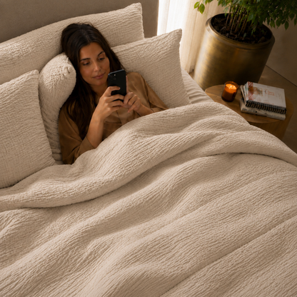 Vrouw liggend in bed onder zandkleurig micro mousseline dekbed zonder overtrek in sfeervolle slaapkamer met kaars