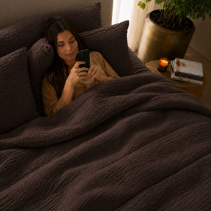 Vrouw liggend in bed onder donkerbruin micro mousseline dekbed zonder overtrek in sfeervolle slaapkamer met kaars
