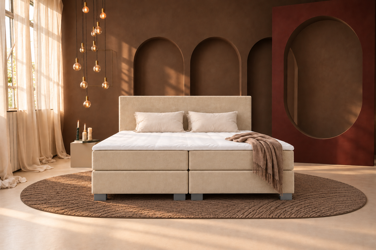 Vooraanzicht van een beige boxspring met gestoffeerd hoofdbord, twee matrassen en een witte topper tegen een mokkabruine muur.